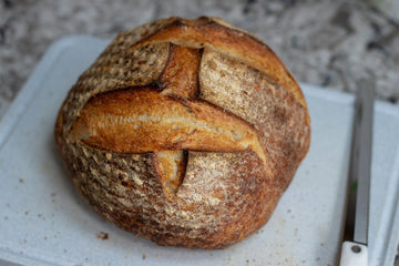 Pan de masa madre redondo y dorado sobre una tabla de cortar, con la corteza rústica marcada en forma de cruz.