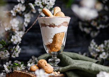 Un tiramisú de kéfir servido en una copa de cristal, decorado con bizcochos y rodeado de flores blancas.