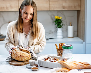 Mujer sonriente preparando pan de masa madre en una cocina moderna, con el Fermentador de masa madre Kefirko y pan recién horneado en la mesa.