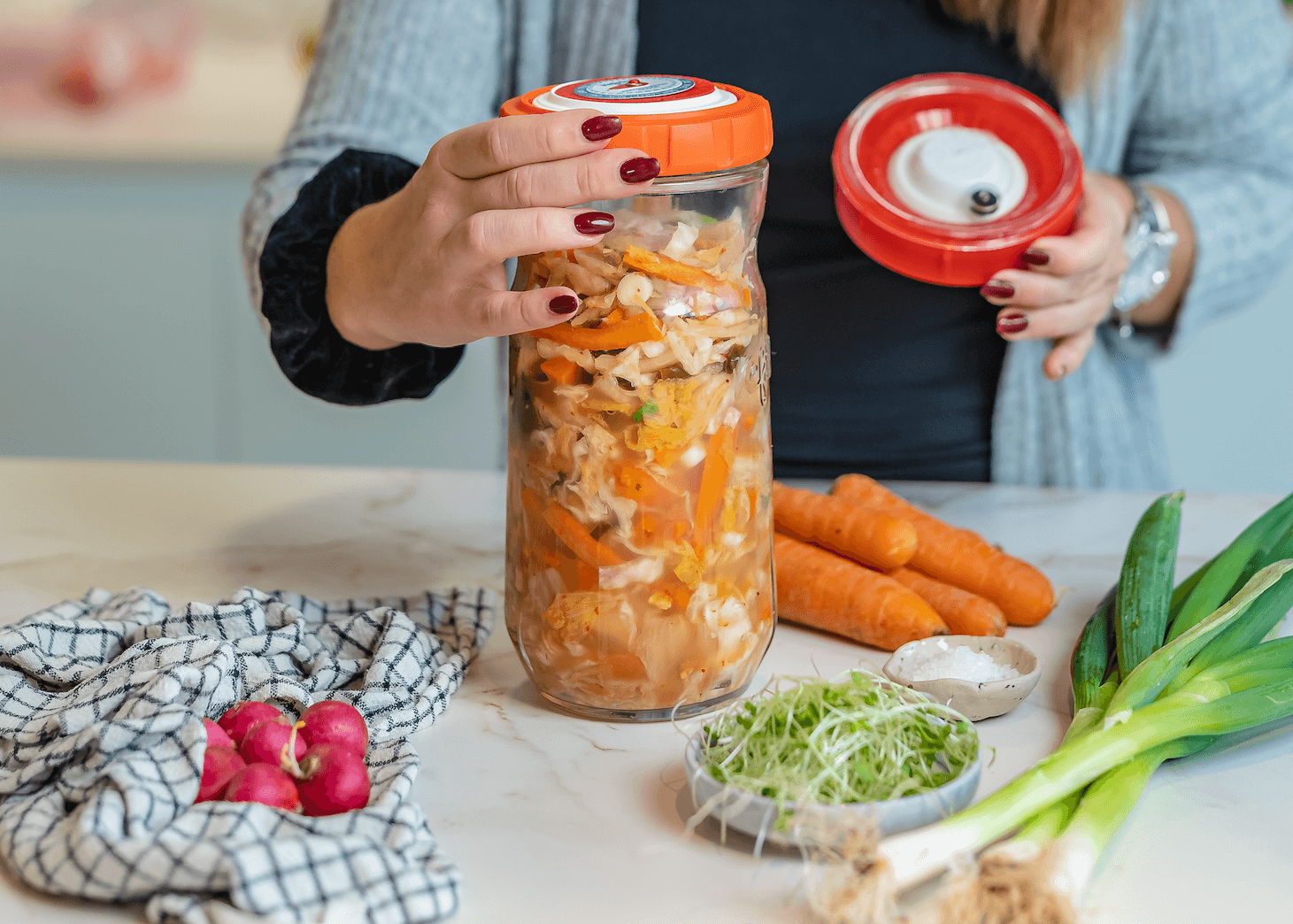 Persona preparando alimentos fermentados en un tarro de vidrio con tapa naranja, rodeado de verduras frescas en una encimera.