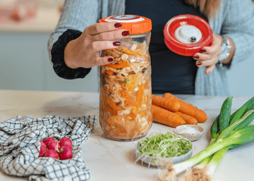 Persona preparando alimentos fermentados en un tarro de vidrio con tapa naranja, rodeado de verduras frescas en una encimera.