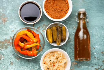 Varios alimentos fermentados como chucrut, pepinillos y pimientos servidos en cuencos sobre una superficie azul.