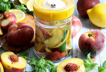 Tarro de vidrio Kefirko con melocotones y limones encurtidos, rodeado de fruta fresca y menta.