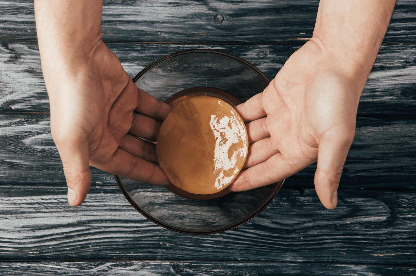 Vista cenital de unas manos sosteniendo un disco de kombucha sobre un bol de cristal en una mesa de madera.