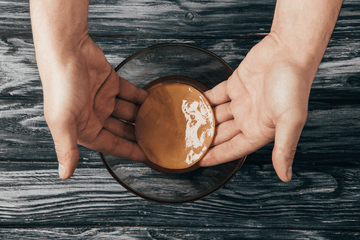 Vista cenital de unas manos sosteniendo un disco de kombucha sobre un bol de cristal en una mesa de madera.