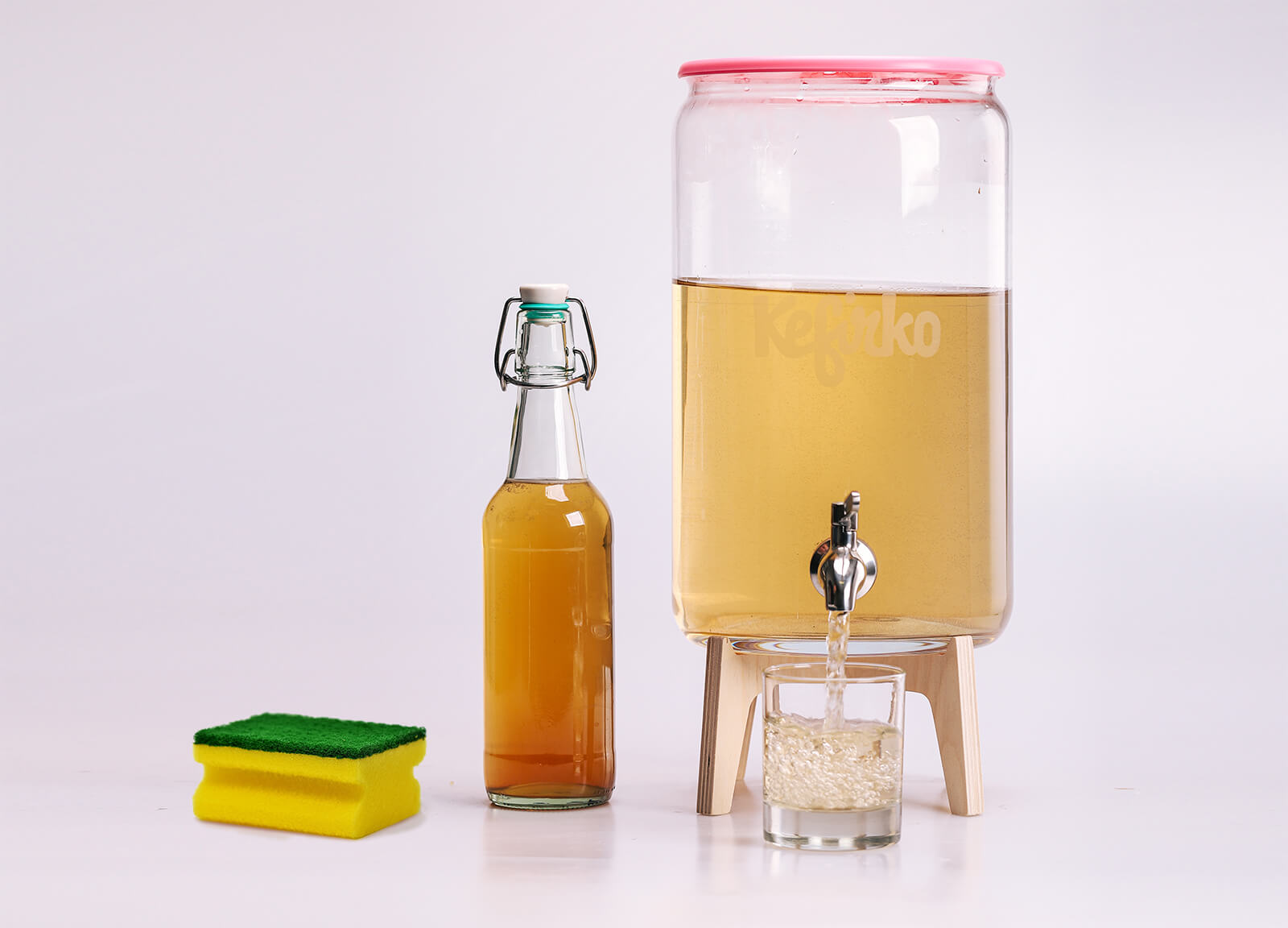 Un tarro de vidrio Kefirko con kombucha, una botella, un vaso y una esponja, listos para preparar un limpiador casero.