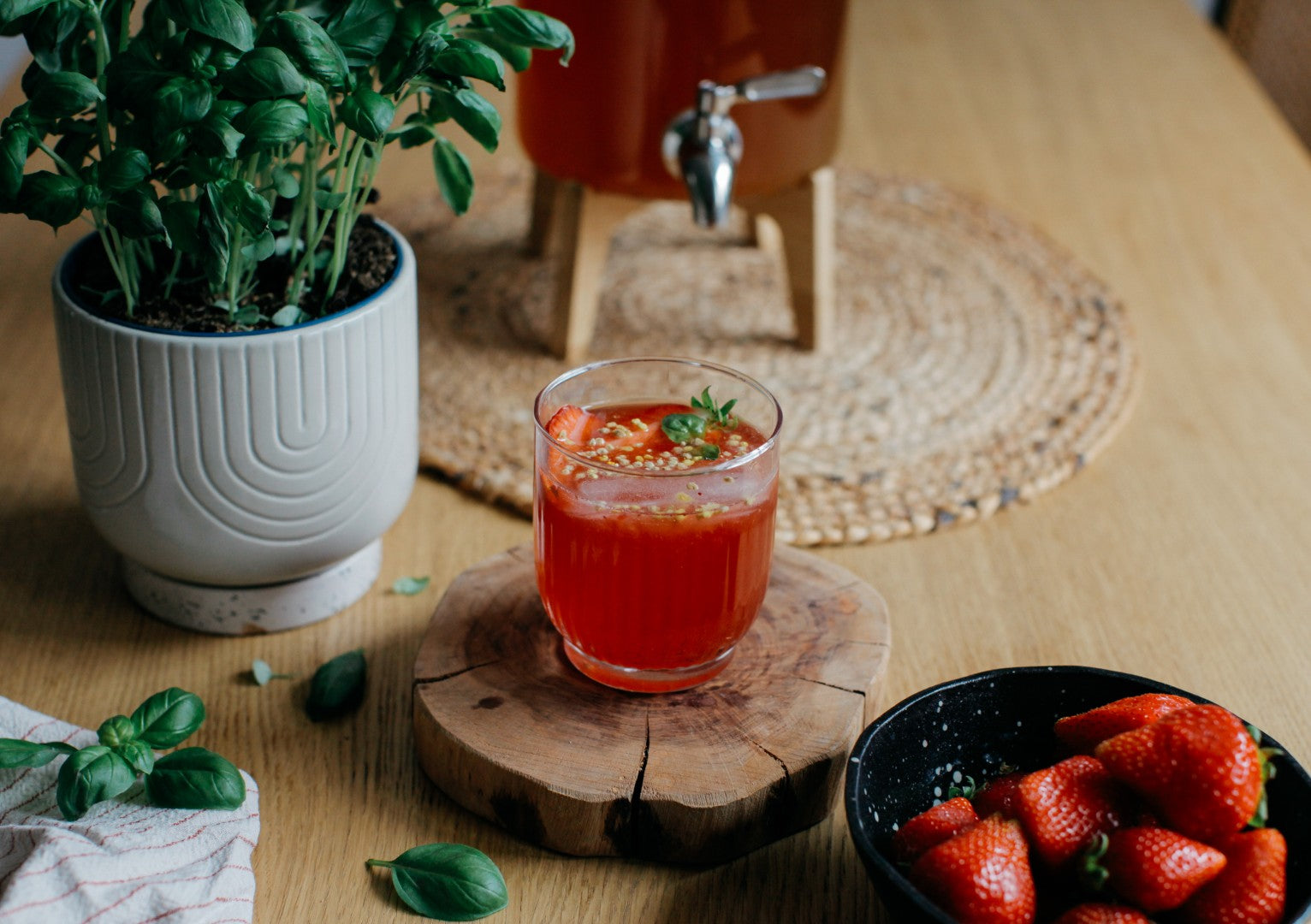 Vaso de cóctel de kombucha con fresas y albahaca sobre un posavasos de madera, junto a un bol de fresas frescas.