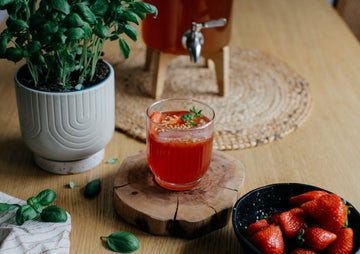 Vaso de cóctel de kombucha con fresas y albahaca sobre un posavasos de madera, junto a un bol de fresas frescas.
