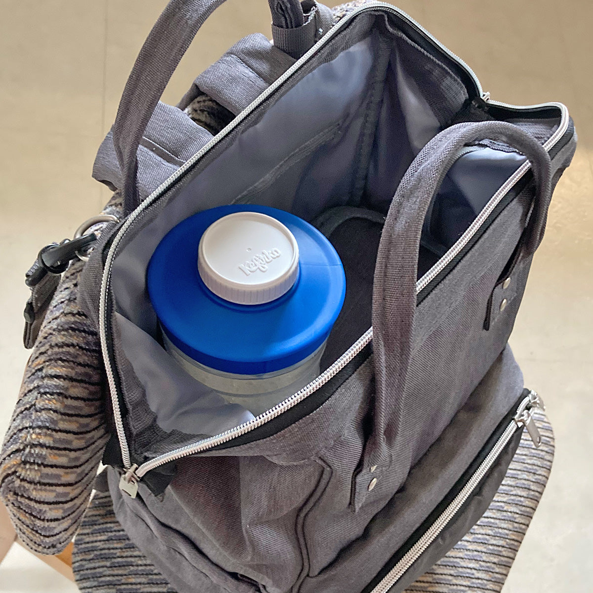 Botella para fermentación Kefirko con su tapa azul y blanca, guardada en el interior de una mochila gris.
