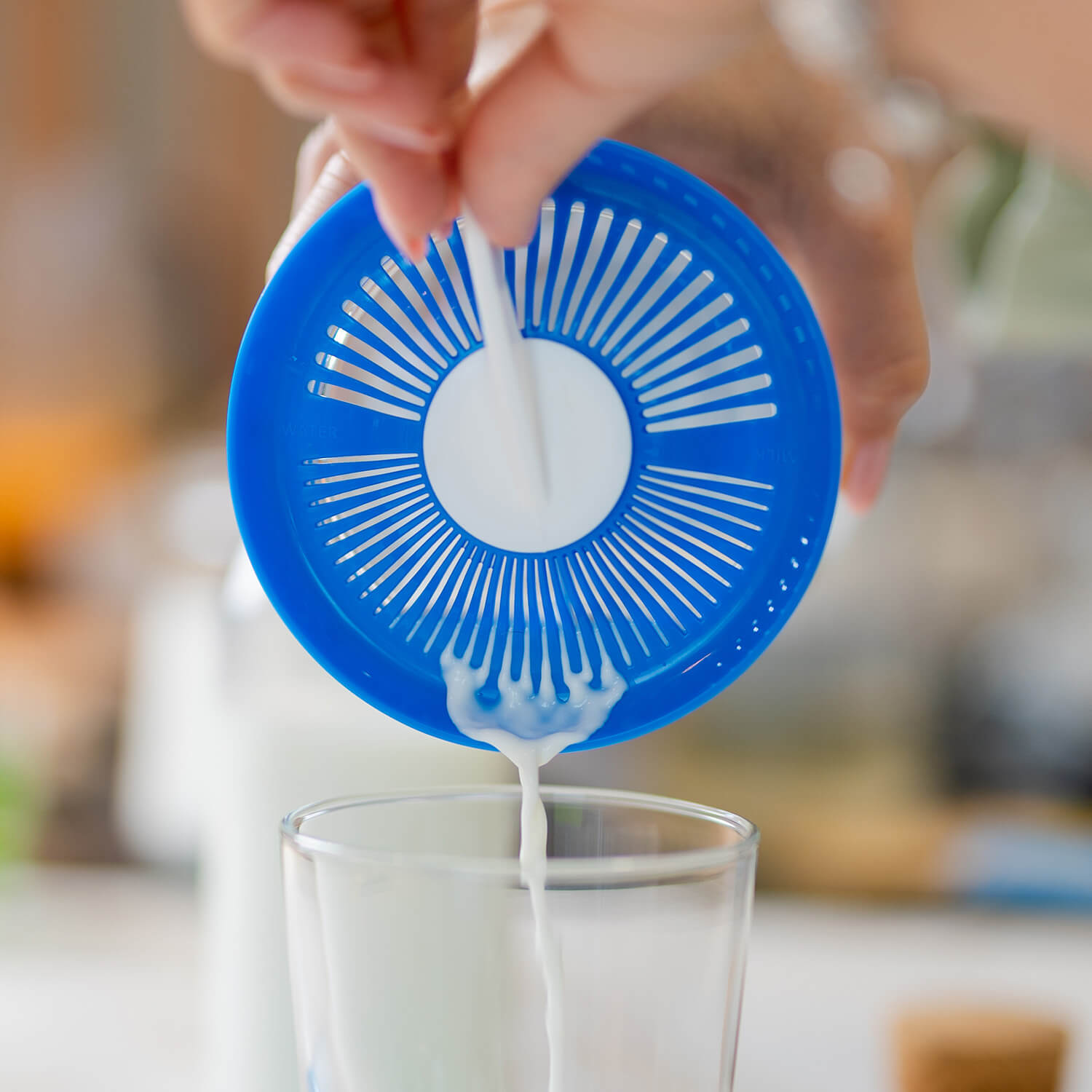 Una mano vierte kéfir de leche desde un tarro, a través de una tapa coladora azul, hacia un vaso de cristal.