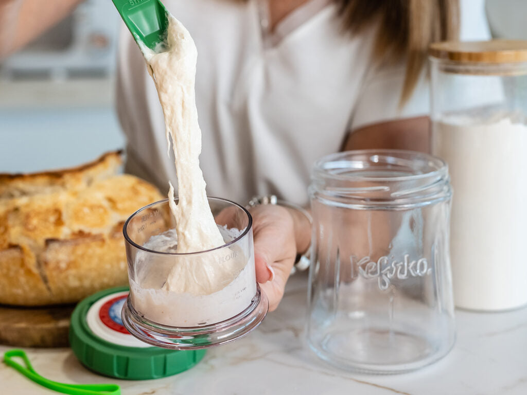 Persona levantando masa madre elástica con una espátula verde del vaso medidor, preparándose para hacer pan de masa madre.