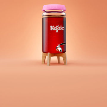 Aquí se muestra un fermentador de vidrio alto Kefirko con tapa rosa y soporte de madera, diseñado para fermentar kombucha. Contiene un líquido rojo y muestra la etiqueta "fermentador de kombucha de 4 l" sobre un fondo naranja.