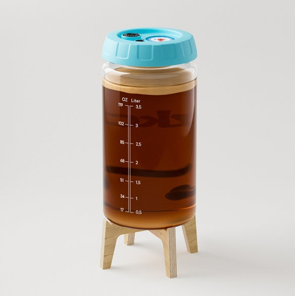 Un recipiente transparente lleno de líquido oscuro, marcado con medidas, se encuentra sobre un pequeño soporte de madera, cubierto con una tapa azul. Es el Fermentador Kombucha 4L de Kefirko en Azul.