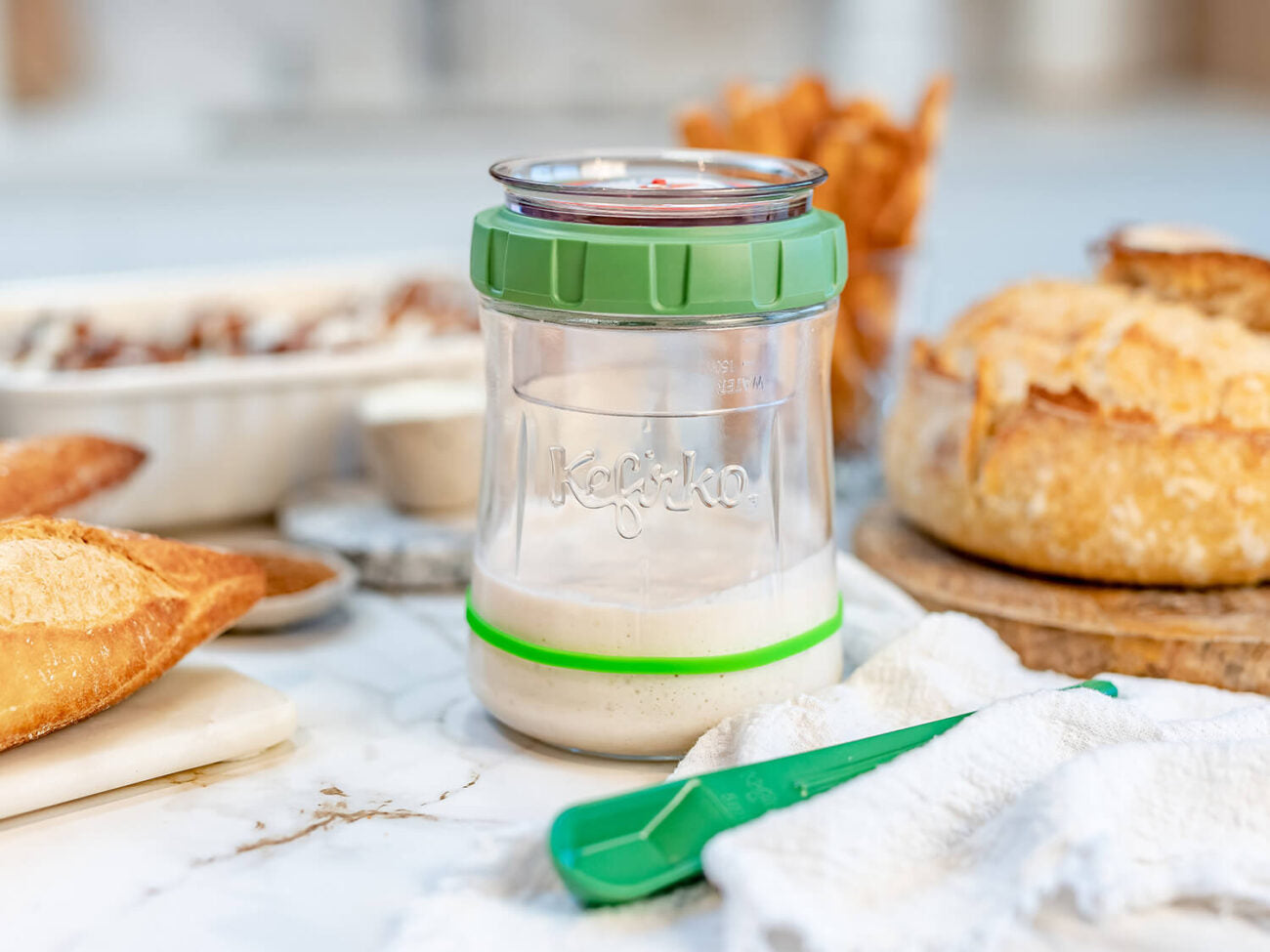 Tarro de vidrio Kefirko con cultivo de masa madre y tapa verde sobre una encimera de mármol junto a pan casero.