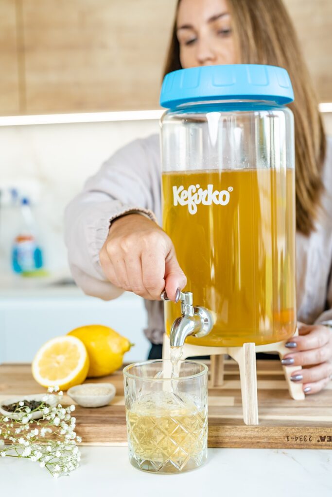 Mujer sirviendo kombucha casera desde el fermentador Kefirko con grifo en un vaso, con limones al lado.