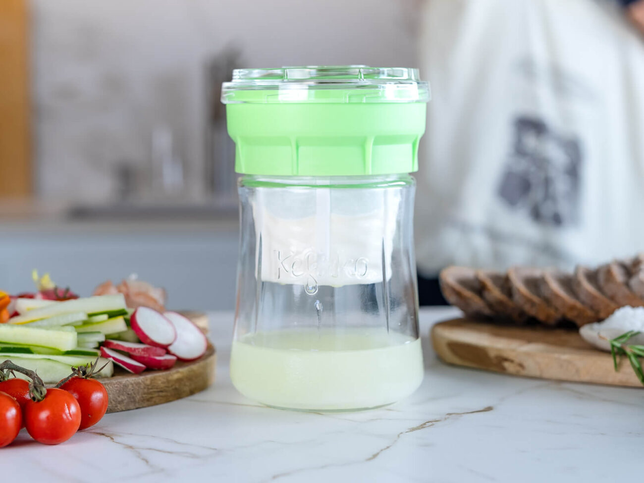 Primer plano del fermentador de queso de Kéfir Kefirko con tapa verde colando suero de kéfir en una cocina con ingredientes frescos.