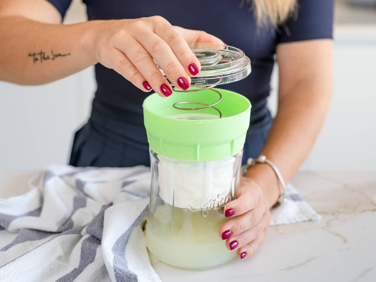 Una mujer abriendo el colador del fermentador de queso de Kéfir Kefirko para mostrar el queso crema recién hecho en una encimera de mármol.