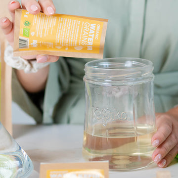 Manos vertiendo el contenido de un sobre en un tarro Kefirko con agua para activar el kéfir.