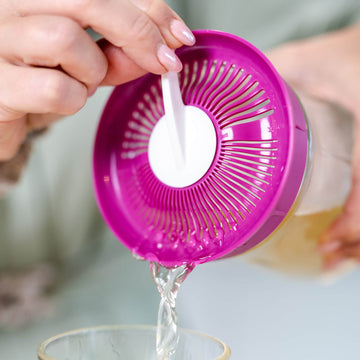 Una persona vierte kéfir de agua desde un tarro con tapa coladora morada a un vaso de cristal.