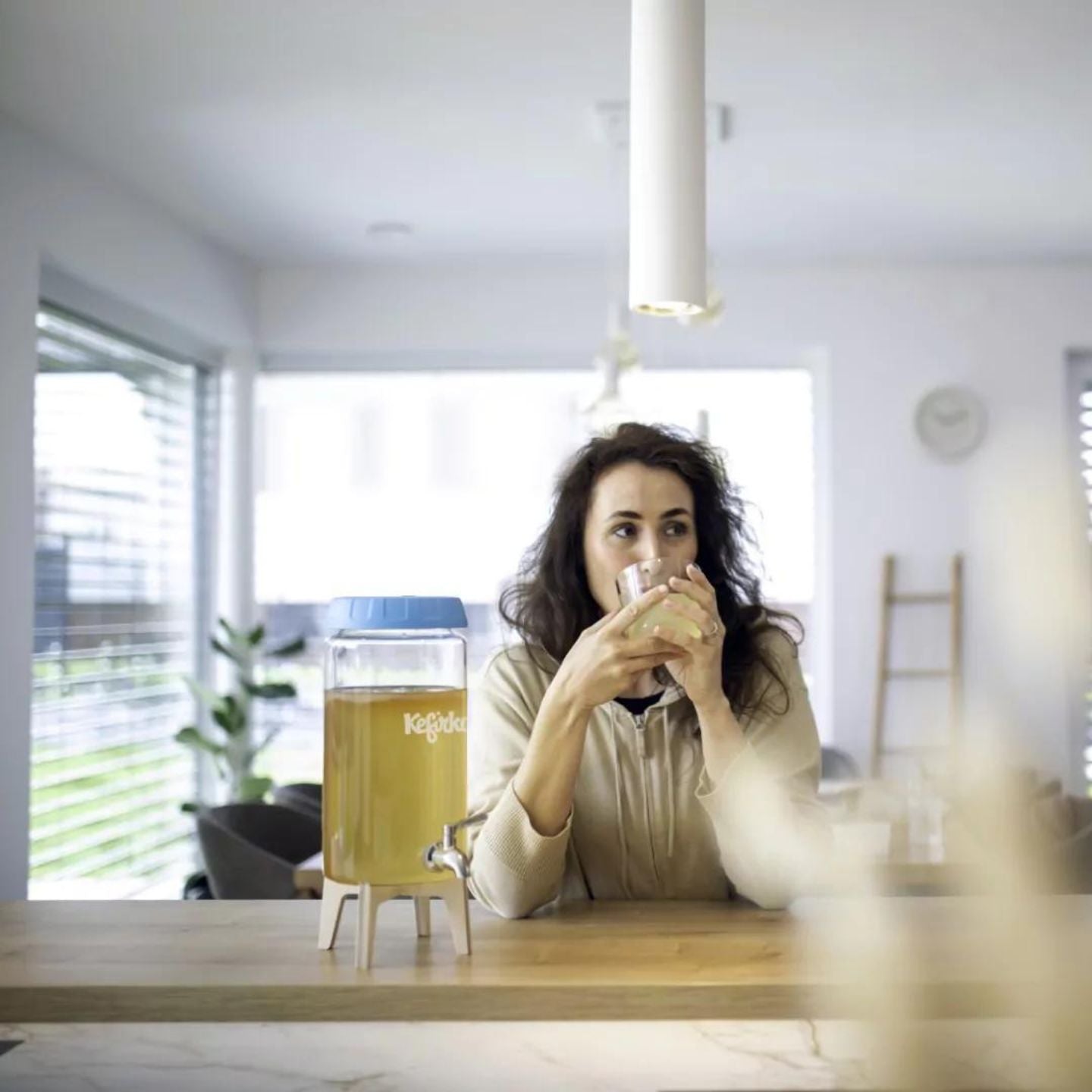 Una persona está sentada en la encimera de la cocina, sosteniendo y bebiendo de un vaso. Hay un dispensador de bebidas grande con la etiqueta "Kefirko" en la encimera. La luz natural ilumina la habitación.