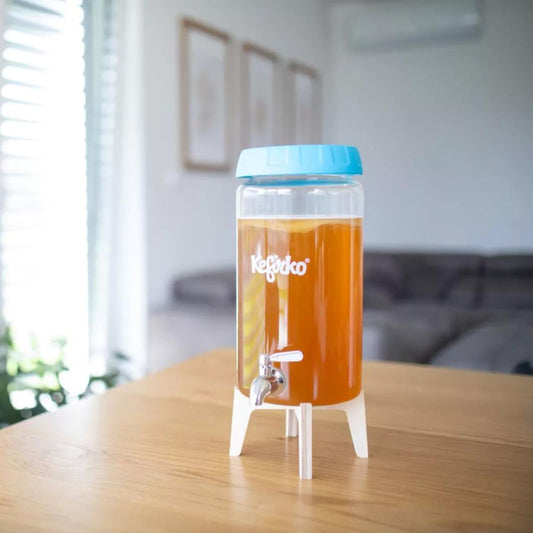 Un dispensador de bebidas Kefirko transparente con tapa azul relleno con kombucha y se coloca sobre una mesa de madera en una habitación luminosa con un sofá al fondo.