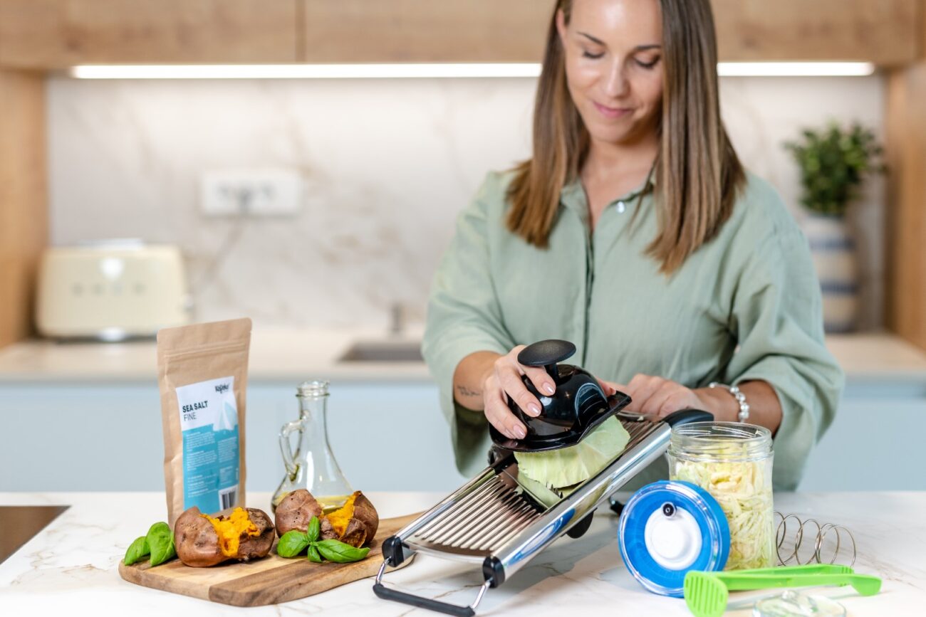Una mujer corta repollo con una mandolina en una cocina, con batatas, especias y utensilios de cocina sobre la encimera. También se ve un recipiente de vidrio lleno de kéfir sobre la encimera.