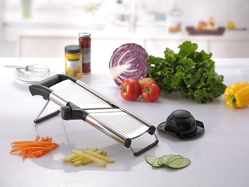 Una mandolina cortadora sobre una encimera de cocina con verduras cortadas en rodajas, lechuga entera, tomates, repollo y pimiento morrón cerca. También hay un recipiente de vidrio lleno de kéfir.