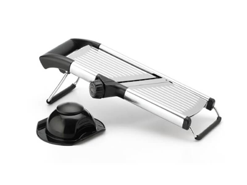 Una mandolina cortadora de acero inoxidable con una cuchilla ajustable y un soporte de seguridad negro a su lado, junto a un recipiente de vidrio lleno de kéfir.