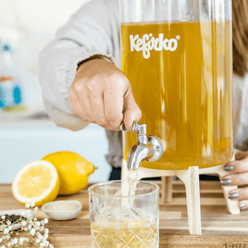Sirviendo kombucha casera desde un dispensador de vidrio Kefirko a un vaso junto a limones.