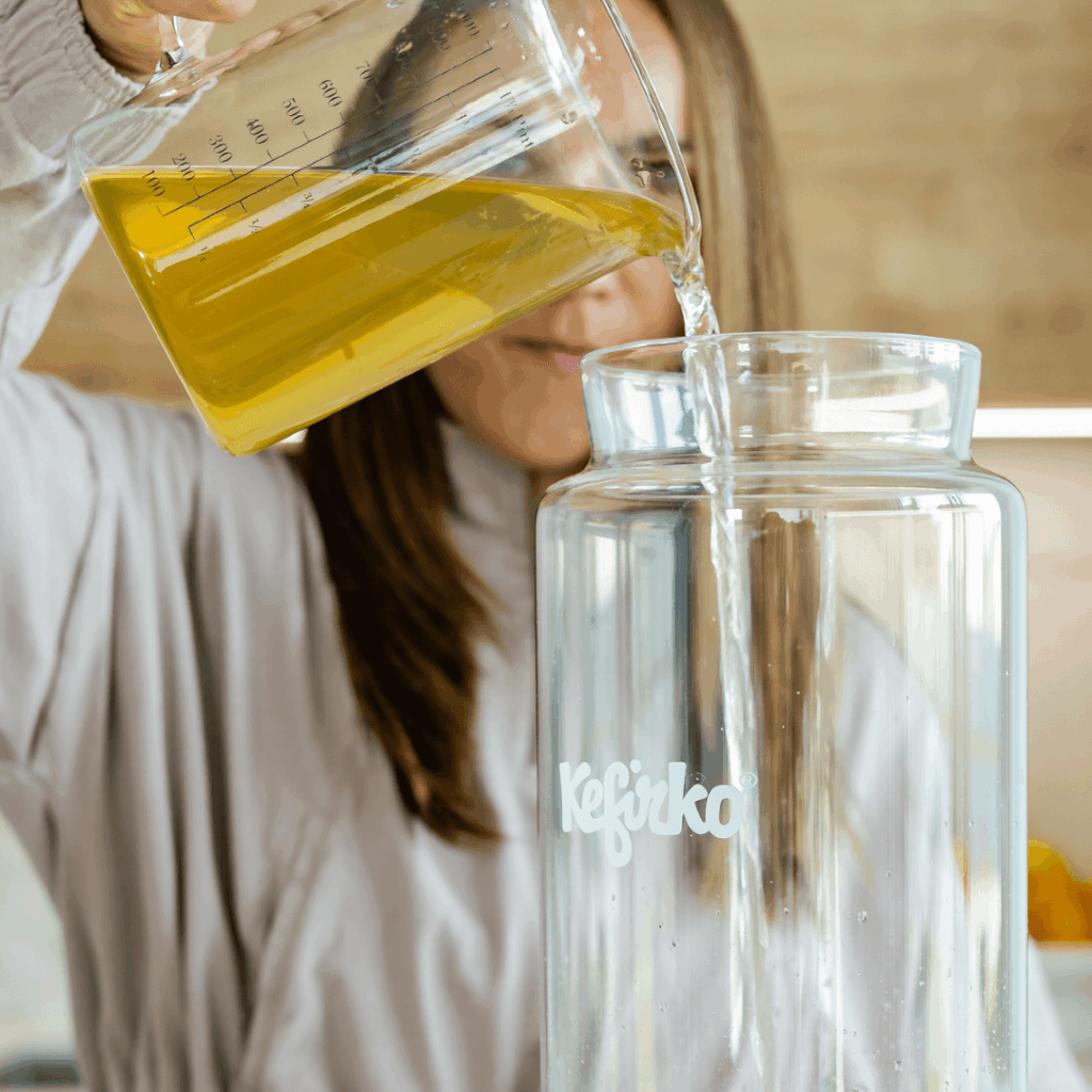 Persona vertiendo té dulce desde una jarra medidora a un tarro de vidrio Kefirko para preparar kombucha.