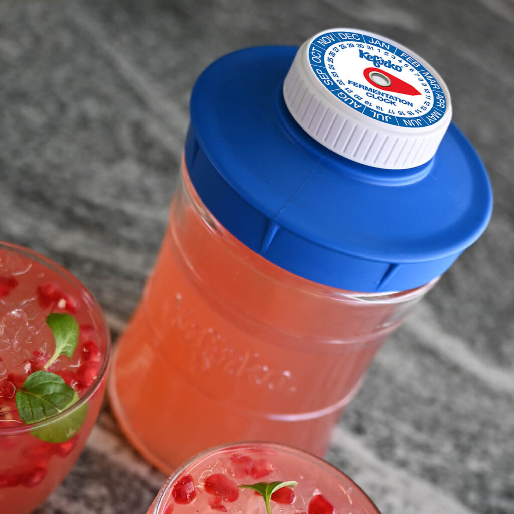 Botella para fermentación con una bebida rosada, junto a dos vasos servidos con kéfir y granada.